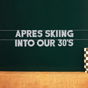 Op de afbeelding: Een zwart schoolbord met de tekst "Apres Skiing into our 30's" geschreven in zilveren glitterletters. De letters zijn aan een lijn met kleine clips bevestigd.