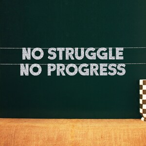 May include: A banner with the words "NO STRUGGLE NO PROGRESS" in silver glitter lettering, suspended against a dark green backdrop. A checkered mug is on the right. The banner is strung on thin white lines.