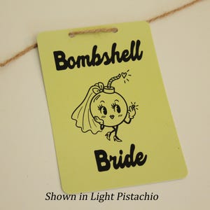Puede incluir: Una etiqueta rectangular de color verde pistacho claro que cuelga de un cordel marrón fino. La etiqueta presenta el texto negro Bombshell Bride y una ilustración de línea negra de una bomba de dibujos animados con velo y anillo.