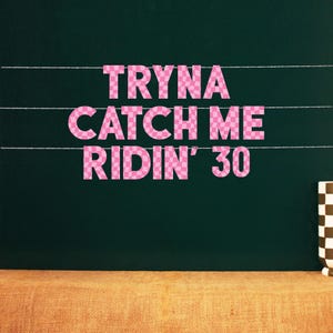 May include: A dark green chalkboard with the text "Tryna Catch Me Ridin' 30" in pink checkered letters. The text is arranged in three lines. White lines are drawn horizontally across the board. A brown and white checkered mug is on the right.