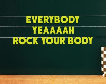 Puede incluir: Una pizarra verde oscuro con las palabras "EVERYBODY YEAAAH ROCK YOUR BODY" en letras de color amarillo brillante. El texto está colgado en el tablero con hilo blanco. Una taza a cuadros blancos y negros está sobre una superficie de arpillera.