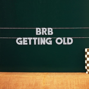 Könnte beinhalten: Eine grüne Tafel mit einem silbernen Glitzerbanner, auf dem "BRB GETTING OLD" steht.