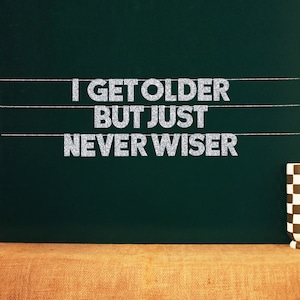 Op de afbeelding: Een groen schoolbord met de tekst "I get older but just never wiser" geschreven in zilveren glitterletters.