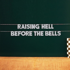 May include: A green chalkboard with the text "Raising Hell Before The Bells" written in silver glitter letters.