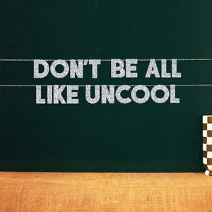 Puede incluir: Una pancarta de purpurina plateada con el texto "Don't be all like uncool" colgada de una pizarra verde oscura.