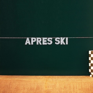 Op de afbeelding: Een zilveren glitterbanner met de tekst "APRES SKI" hangt aan een donkergroen schoolbord.