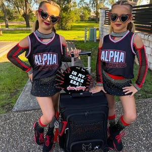 Può includere: Due ragazze giovani che indossano uniformi da cheerleader nere e rosse con la scritta "ALPHA" sul davanti. Stanno tenendo una valigia nera con rotelle con un'immagine in bianco e nero di un gatto sul davanti. La valigia ha un cartello in bianco e nero che dice "IN MY CHEER ERA cheer-sisters".