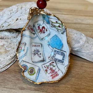 Op de afbeelding: Een oesterschelp ornament met een goud geschilderde rand, met een handgeschilderd ontwerp van Mahjong tegels. Het ornament wordt opgehangen door een gouden koord met een rode en gouden kraal. De schelp staat tegen een houten achtergrond.