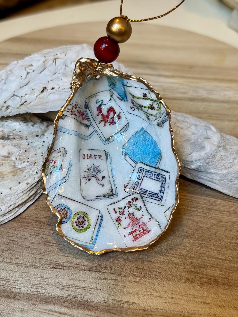 Op de afbeelding: Een oesterschelp ornament met een goud geschilderde rand, met een handgeschilderd ontwerp van Mahjong tegels. Het ornament wordt opgehangen door een gouden koord met een rode en gouden kraal. De schelp staat tegen een houten achtergrond.