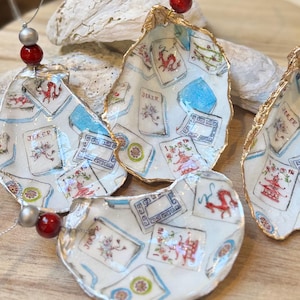 Op de afbeelding: Handbeschilderde oesterschelp ornamenten met speelkaarten ontwerpen in rood, blauw en goud. Elke schelp is uniek, geregen met kralen en toont een 'Joker' kaart. Geschikt voor huisdecoratie.