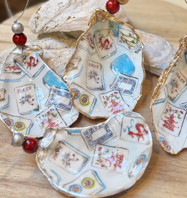 Op de afbeelding: Handbeschilderde oesterschelp ornamenten met speelkaarten ontwerpen in rood, blauw en goud. Elke schelp is uniek, geregen met kralen en toont een 'Joker' kaart. Geschikt voor huisdecoratie.