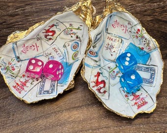 Op de afbeelding: Twee decoratieve oesterschelpen met goudkleurige randen, voorzien van decoupage-illustraties van mahjongstenen en speelkaarten. De linker schelp bevat twee doorschijnende roze dobbelstenen, de rechter schelp twee doorschijnende blauwe dobbelstenen.