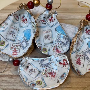 Op de afbeelding: Vier oesterschelp ornamenten met een gouden rand. De schelpen zijn versierd met een witte achtergrond en een patroon van speelkaarten, pagodes en draken. Elk ornament heeft een kralenketting om op te hangen.