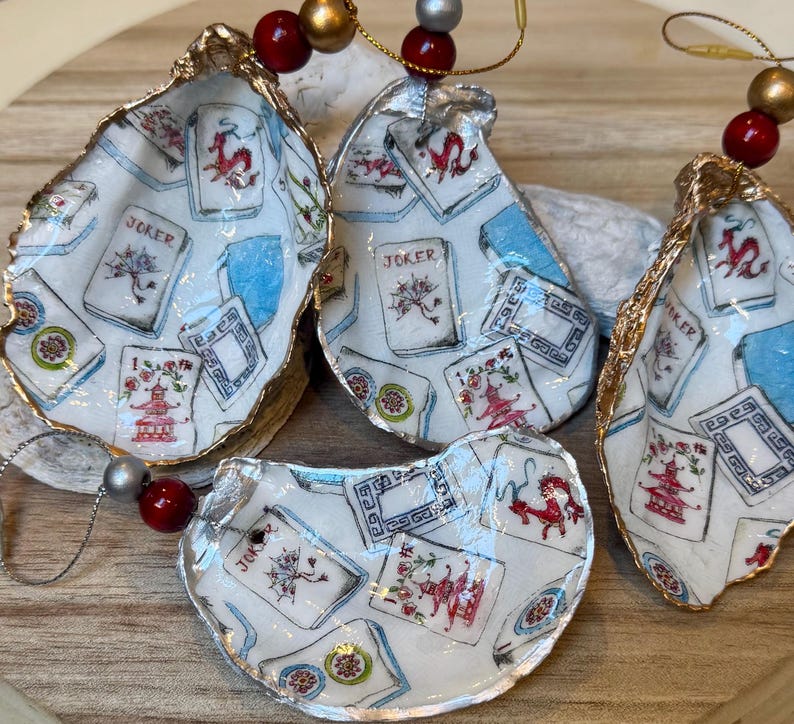 Op de afbeelding: Vier oesterschelp ornamenten met een gouden rand. De schelpen zijn versierd met een witte achtergrond en een patroon van speelkaarten, pagodes en draken. Elk ornament heeft een kralenketting om op te hangen.