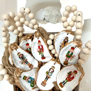 May include: A set of eight oyster shell ornaments featuring hand-painted nutcracker designs. Each ornament is painted with a different nutcracker design, including a red and gold nutcracker, a blue and green nutcracker, and a gold and white nutcracker. The ornaments are displayed in a white box with a gold trim.