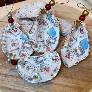 Op de afbeelding: Vier oesterschelp ornamenten met speelkaarten ontwerpen. De schelpen zijn beschilderd met speelkaartenmotieven, waaronder "Joker" kaarten, en hebben goudkleurige randen. Elk ornament is geregen met kralen.