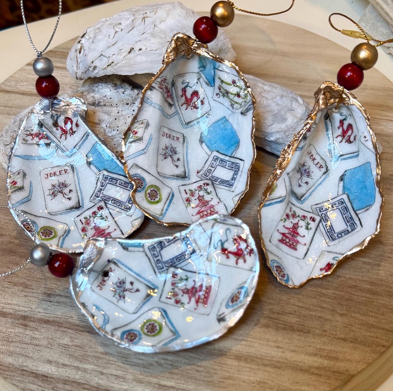 Op de afbeelding: Vier oesterschelp ornamenten met speelkaarten ontwerpen. De schelpen zijn beschilderd met speelkaartenmotieven, waaronder "Joker" kaarten, en hebben goudkleurige randen. Elk ornament is geregen met kralen.