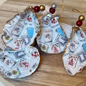 Op de afbeelding: Vier oesterschelp ornamenten met een speelkaarten ontwerp. De schelpen hebben een gouden rand en zijn versierd met speelkaarten afbeeldingen, inclusief het woord "Joker". Elk ornament is geregen met kralen en goudkleurig draad.