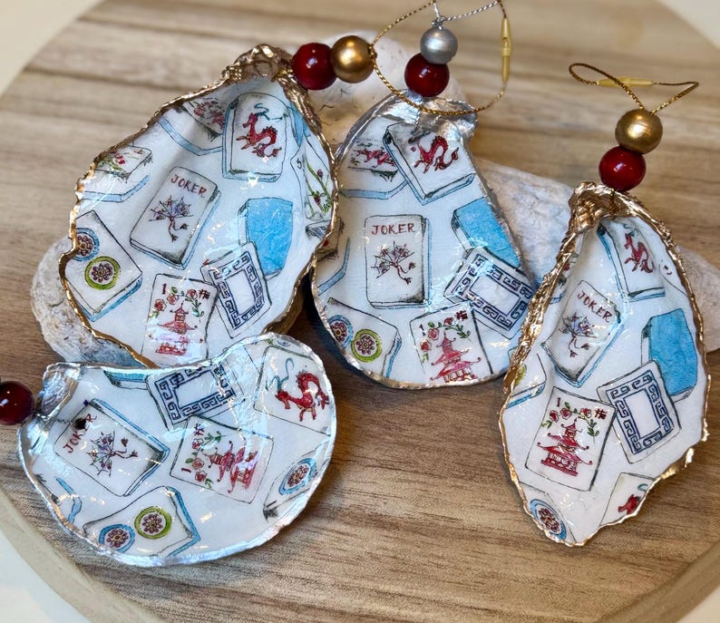 Op de afbeelding: Vier oesterschelp ornamenten met een speelkaarten ontwerp. De schelpen hebben een gouden rand en zijn versierd met speelkaarten afbeeldingen, inclusief het woord "Joker". Elk ornament is geregen met kralen en goudkleurig draad.