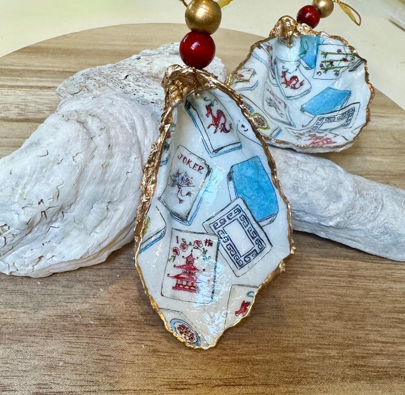 Op de afbeelding: Handbeschilderde oesterschelpversieringen met gouden rand, met speelkaartenmotieven. De schelpen zijn versierd met rode en gouden kralen om op te hangen. Deze ornamenten bieden een unieke, artistieke stijl, ideaal voor feestelijke decoratie of als een onderscheidend cadeau.