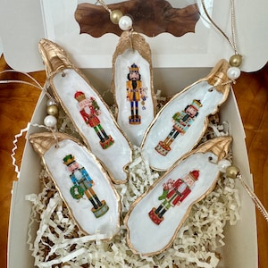 May include: Set of five gold-painted oyster shell ornaments with nutcracker designs. Each ornament features a different nutcracker with a festive holiday theme. The ornaments are packaged in a white box with gold trim and white tissue paper.