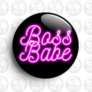 Może przedstawiać: Czarny guzik z napisem "Boss Babe" w jaskraworóżowym neonowym stylu. Guzik ma błyszczące wykończenie i jest umieszczony na tle powtarzających się białych i szarych grafik z dymkami.