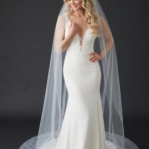 Puede incluir: Vestido de novia blanco con escote pronunciado y delicados detalles de encaje. El vestido tiene una silueta ajustada que se ensancha en la parte inferior. Un velo transparente y fluido cae sobre los hombros, creando un look nupcial elegante.