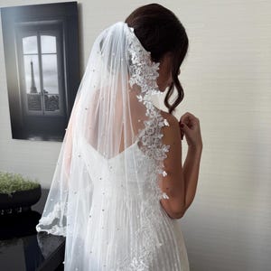 Puede incluir: Un velo de novia blanco con delicados bordes de encaje y acentos de perlas dispersas. El velo está drapeado sobre un vestido de novia blanco, creando un aspecto suave y romántico. El encaje presenta diseños florales.