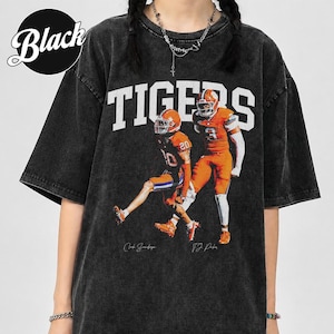 May include: A black acid-wash short-sleeve t-shirt featuring the word TIGERS in large white letters above a graphic of two football players in orange uniforms. The players are numbered 10 and 3, with signatures printed below each figure.
