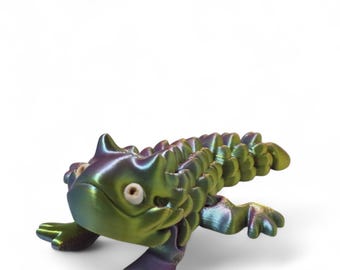 May include: A small, green, iridescent, 3D printed figurine of a horned lizard. The lizard has a spiky back and a wide, flat head with large eyes.