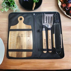 Peut inclure: Une mallette de transport noire contenant une planche à découper en bois et trois outils de grillade. La planche à découper porte l'inscription "BBQ TIME" et "ORDER PIZZA". Les outils comprennent une spatule, des pinces et une brosse à grillade.