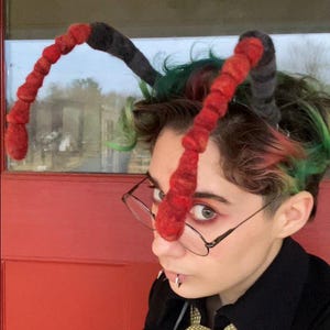 May include: A person with green and pink hair is wearing a black jacket, a gray vest, and a green patterned tie. They are wearing a pair of glasses and have a red and black felt ant antennae headband on their head.