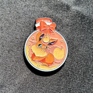 May include: A round metal enamel pin featuring a stylized orange fox-like creature with large blue eyes and a cream-colored chest tuft, enclosed within a circular bottle shape topped with a red bow and a white cap, set against a soft orange background.