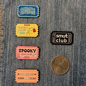 May include: Five rectangular enamel pins shaped like admission tickets. The pins feature text including Read All Night Book Club, Emotional Damage Book Club, Smut Club, Spooky Book Club, and Romance Book Club. A US quarter coin is shown for scale.