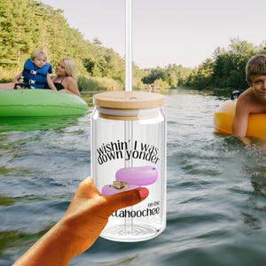 May include: A clear glass tumbler with a bamboo lid and a straw, held in a hand. The tumbler has the text "wishin' I was down yonder on the catahoochee" and a graphic of a pink inner tube. The background shows people floating in a river.