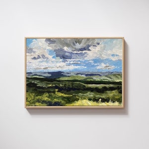 May include: An original landscape painting featuring a vibrant sky with white and gray clouds over rolling green hills. The artwork is framed in a natural wood frame, creating a serene and natural aesthetic.