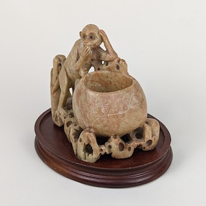 Puede incluir: Una escultura de piedra tallada de un mono que se tapa las orejas, sentado junto a una olla redonda de piedra tallada. La escultura está sobre una base de madera.