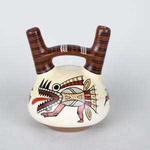 May include: A cream-colored ceramic vessel with a brown handle and spouts. The vessel is decorated with a stylized creature design in red, brown, and beige. The creature has an open mouth and a long red tongue.