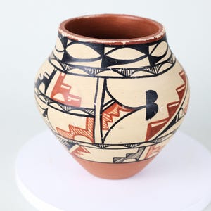 Puede incluir: Una olla de cerámica pintada a mano con un cuerpo redondeado y una abertura ancha. La olla presenta un fondo color crema con patrones geométricos en negro y marrón rojizo. El borde y la base son de color terracota.