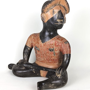 Pode incluir: Uma estatueta sentada, esculpida à mão, com corpo preto e rosto e chapéu bege. A estatueta usa uma camisa vermelha com um padrão geométrico. A figura está segurando um objeto de cor escura.