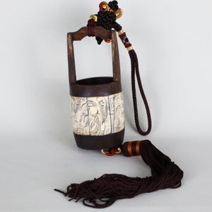 May include: A cylindrical wooden bucket with a dark brown finish and a central cream-colored panel featuring black line drawings of figures. The bucket has a dark brown handle, a braided cord, and a long dark brown tassel hanging from the base.