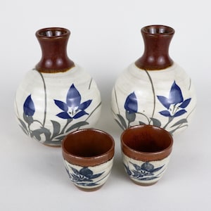 May include: A ceramic sake set featuring two round, white and brown bottles with dark brown necks and two matching cups. Each piece is decorated with blue floral designs and gray stems.