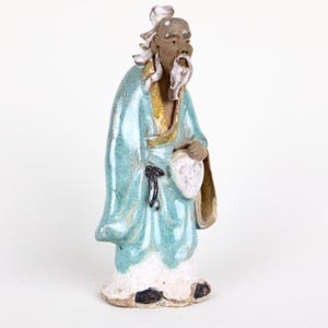 Może przedstawiać: Ceramiczna figurka starszego mężczyzny w powłóczystej szacie. Postać jest głównie błękitna ze złotymi wykończeniami i białą brodą. Trzyma przedmiot w kształcie wachlarza i ma spokojny wyraz twarzy. Figurka jest prawdopodobnie pochodzenia azjatyckiego.