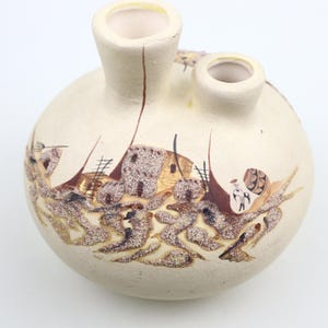 Peut inclure: Un vase en céramique blanche avec deux ouvertures, peint à la main avec une scène de maisons et de personnages amérindiens. Le vase a une surface texturée et des accents marrons.