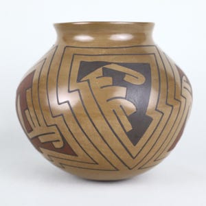 May include: A tan-colored ceramic pot with a rounded body and a slightly flared rim. The pot features geometric designs in black and reddish-brown, creating a striking contrast against the tan background. The design includes angular shapes and stylized motifs.