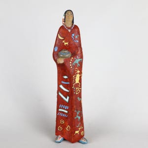 May include: A tall, slender figurine of a person with dark hair, wearing a long, textured red robe adorned with various painted symbols including a yellow crescent moon, a goat, a lizard, and geometric shapes, while holding a small gray bowl.