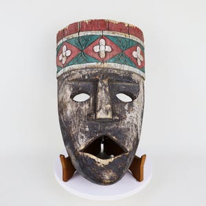 May include: A weathered wooden mask with a distressed finish. The mask features a red, green, and white floral patterned band across the top. The mask is mounted on a white circular base with a wooden stand.