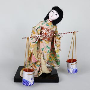 May include: A Japanese doll in a traditional kimono, carrying two buckets suspended from a pole. The kimono is pink with floral patterns. The buckets are silver with blue wave designs. The doll is on a black base.