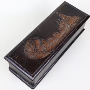 May include: A dark brown rectangular wooden box featuring a carved relief on the lid. The relief shows a detailed scene with figures and buildings. The box has a glossy finish and appears to be vintage, possibly antique.