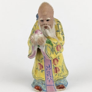 May include: A ceramic figurine of an elderly man with a long white beard, wearing a yellow robe with red floral patterns. The figurine holds a pink object and has a blue accent on the back.